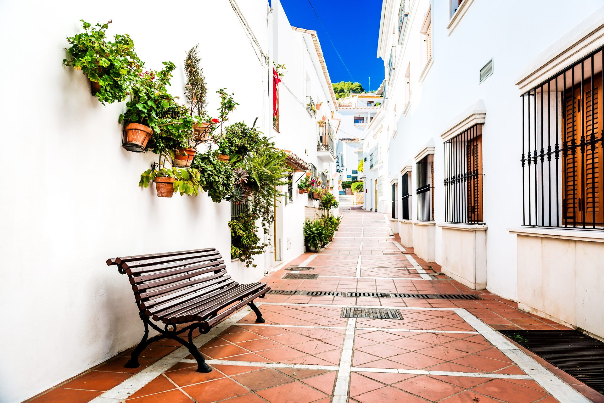 Narrow alley with white walls, potted plants, wooden bench, and blue sky.